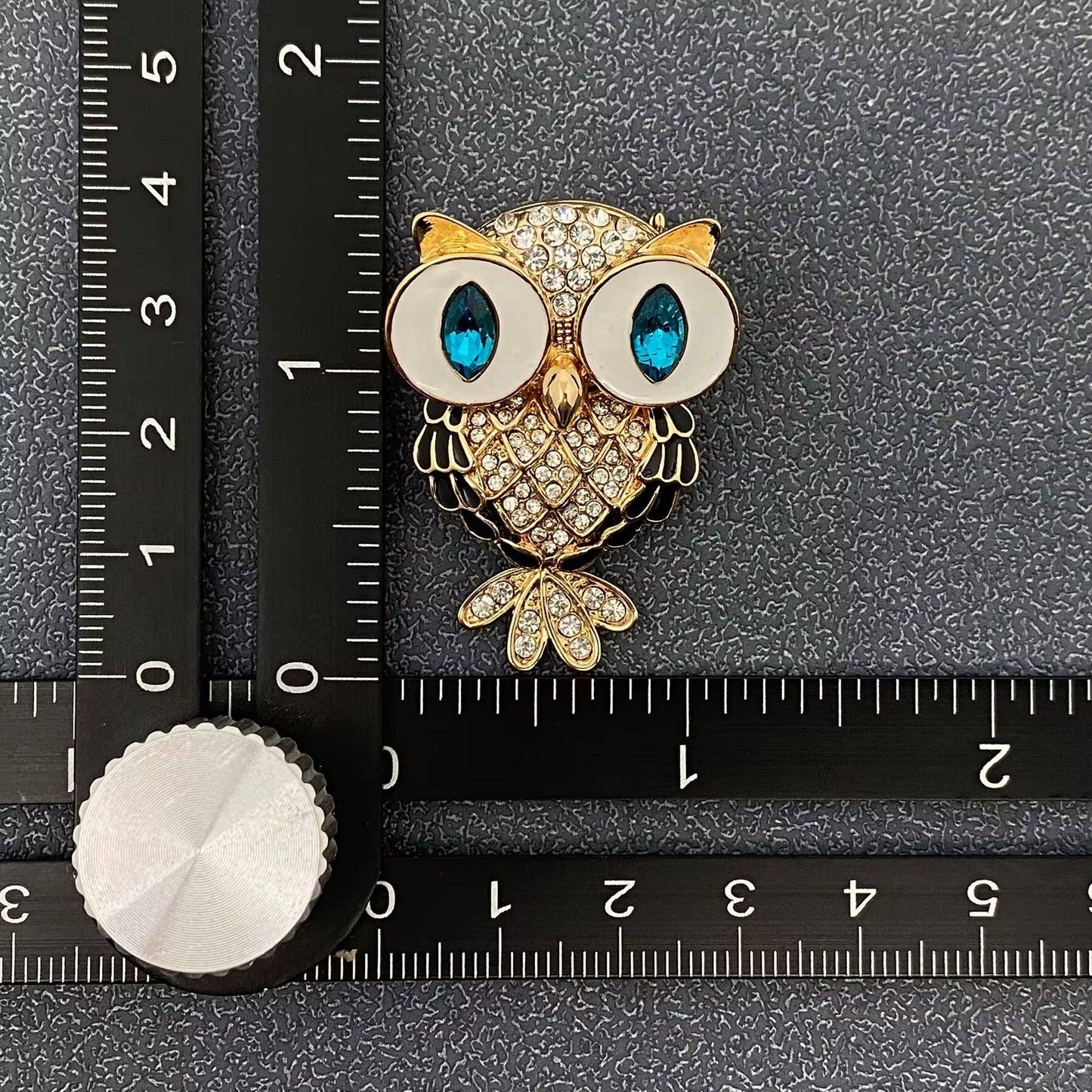 Gold owl brooch with blue eyes on a black surface with a ruler for scale.