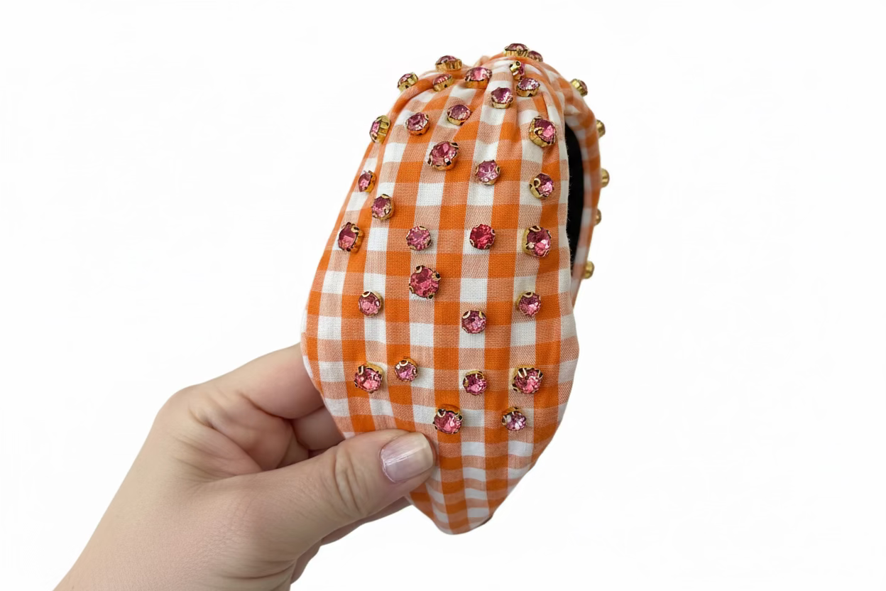 hand holding an orange and white gingham headband with crystal embellishment against a white background