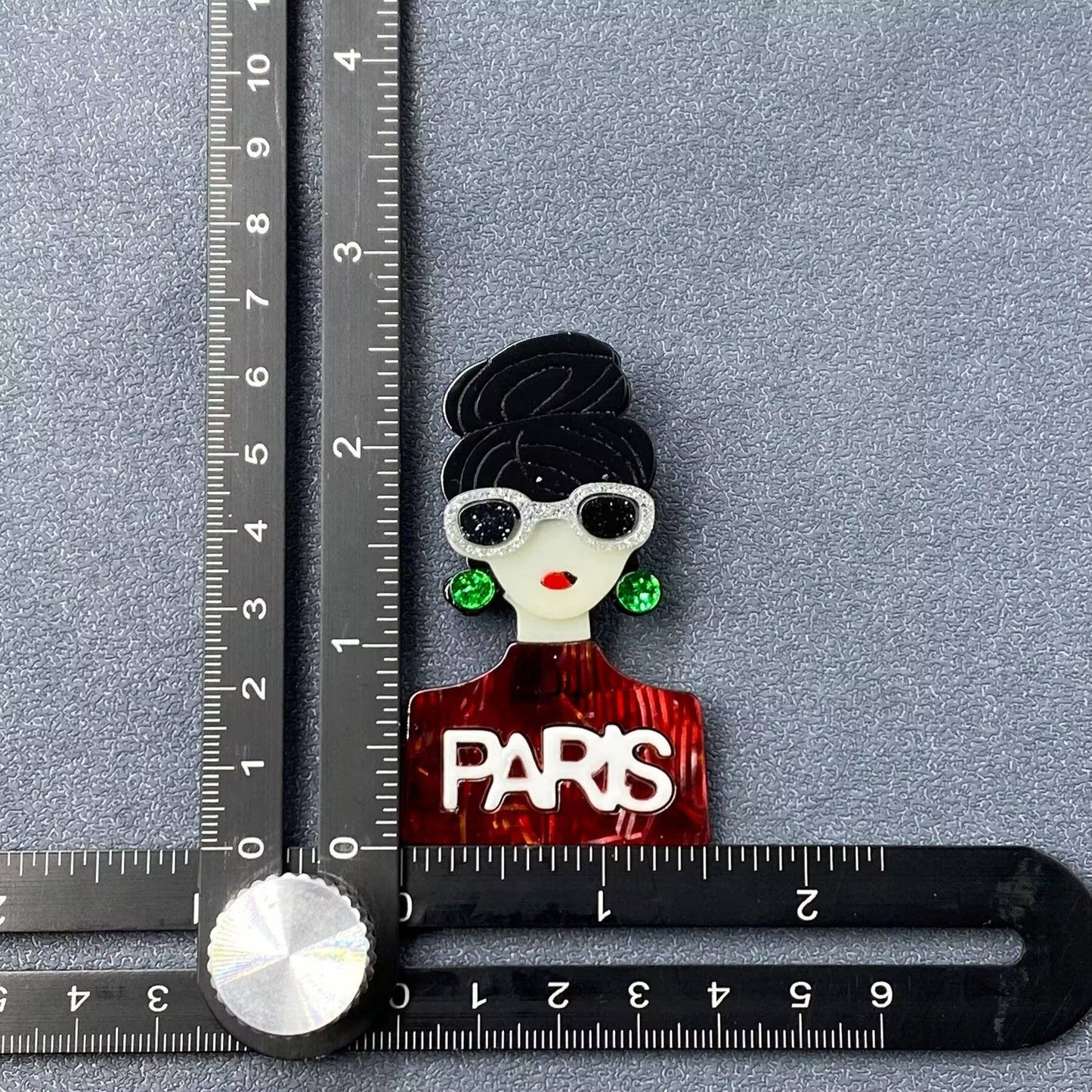 Brooch feautring a woman with 'PARIS' text on a gray background with ruler for scale