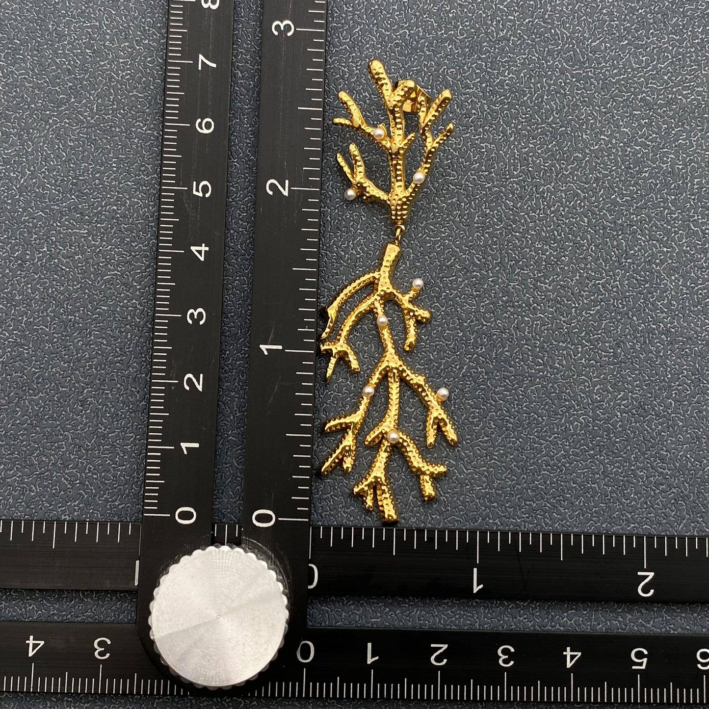 one gold pearl encrusted coral shaped earrings next to a ruler on a grey surface