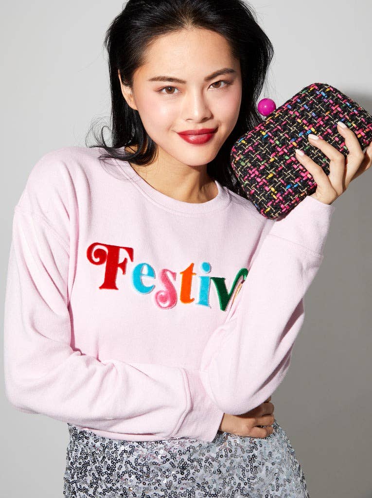 woman wearing a sweatshirt with "Festive" written on the front and holding a black multi colored tweed clutch