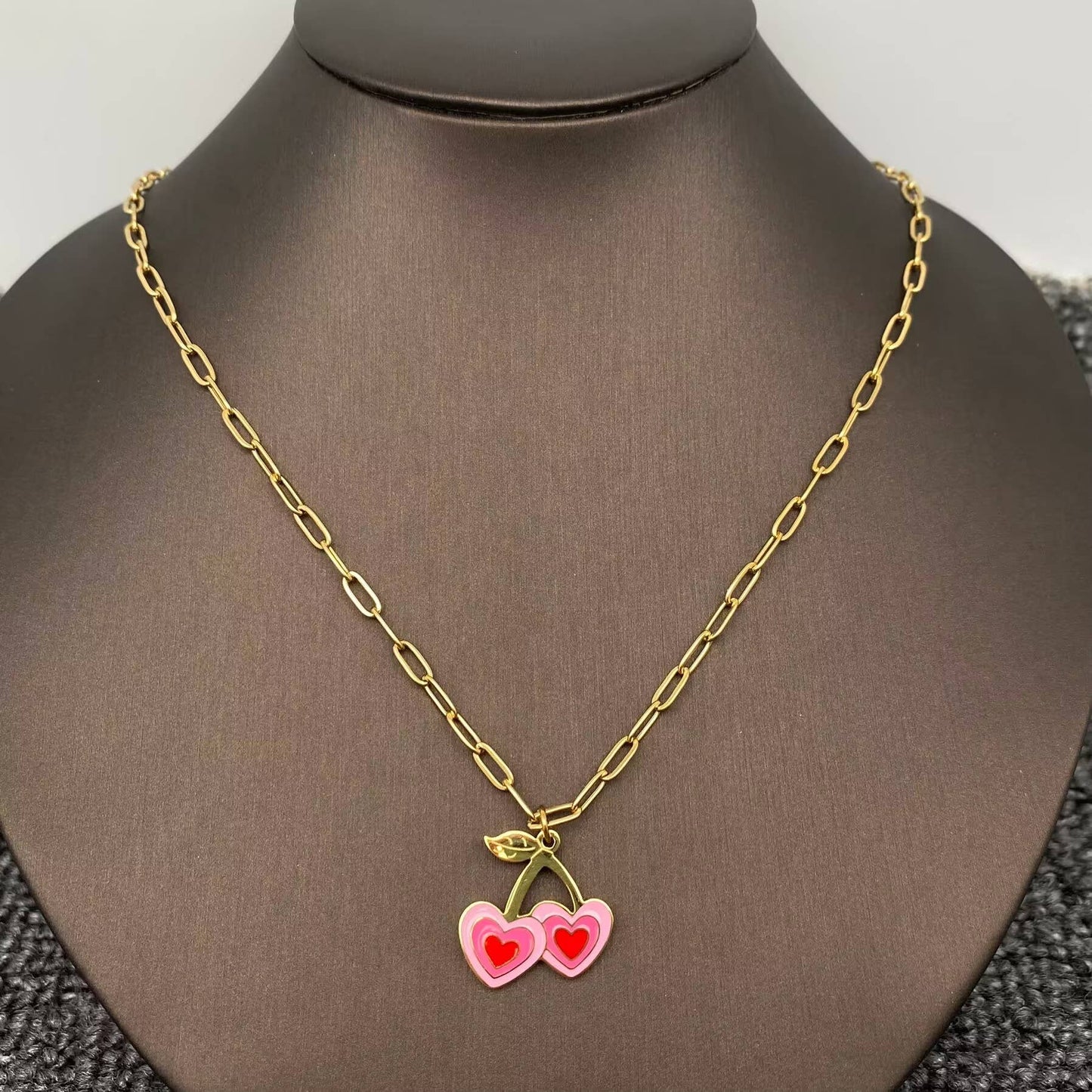 a pink and red cherry themed enamel necklace with gold chain on a black necklace stand