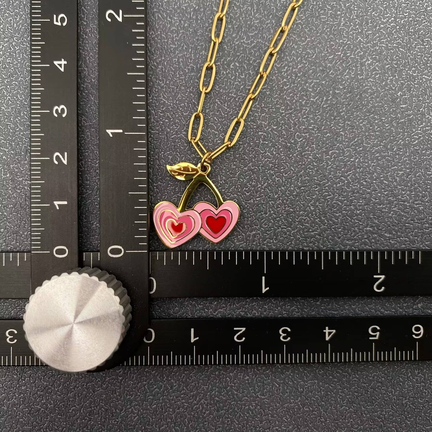 up close view of a pink and red cherry themed pendant necklace with gold chain next to a black ruler laying on a grey background