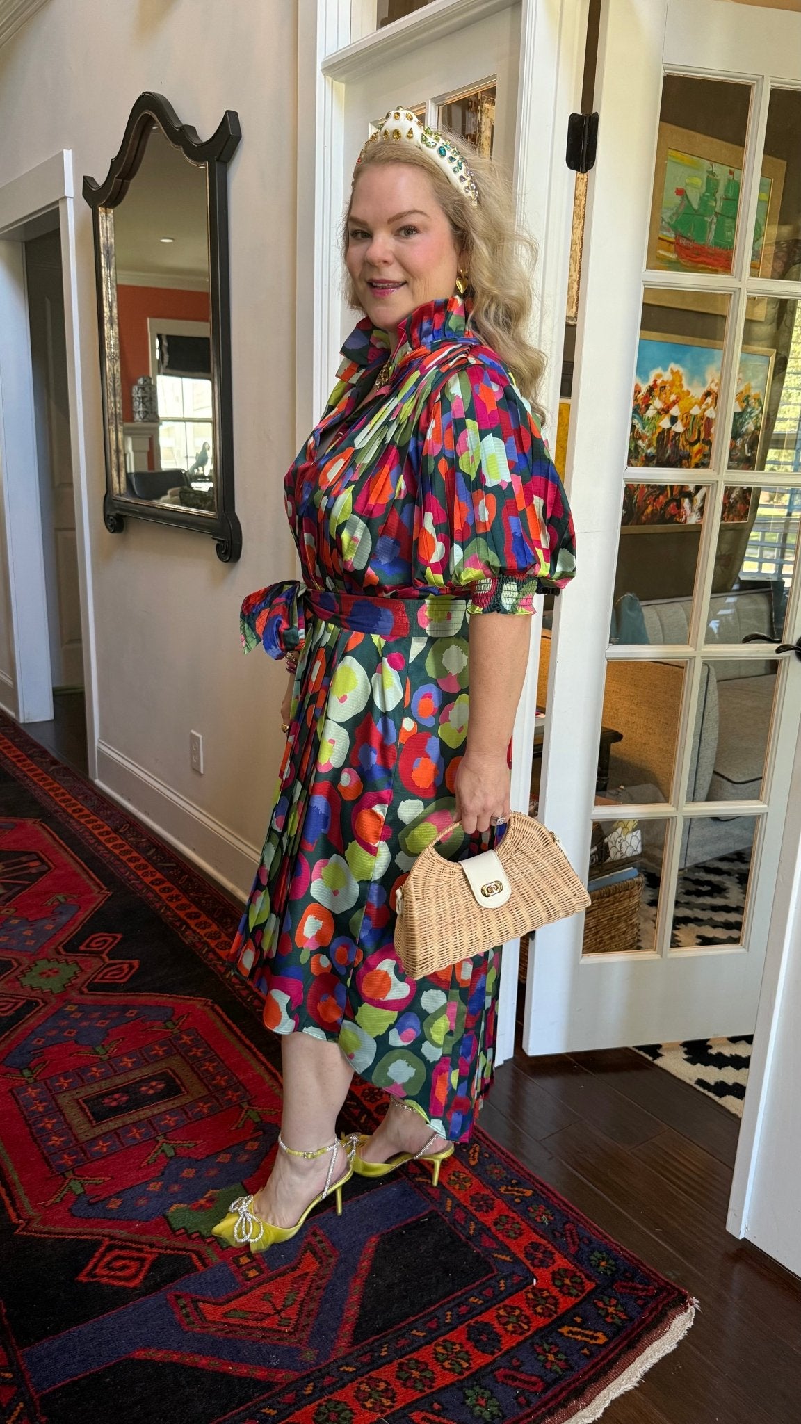 Woman in a colorful dress and wearing a rattan handbag standing in a room with a mirror and door.