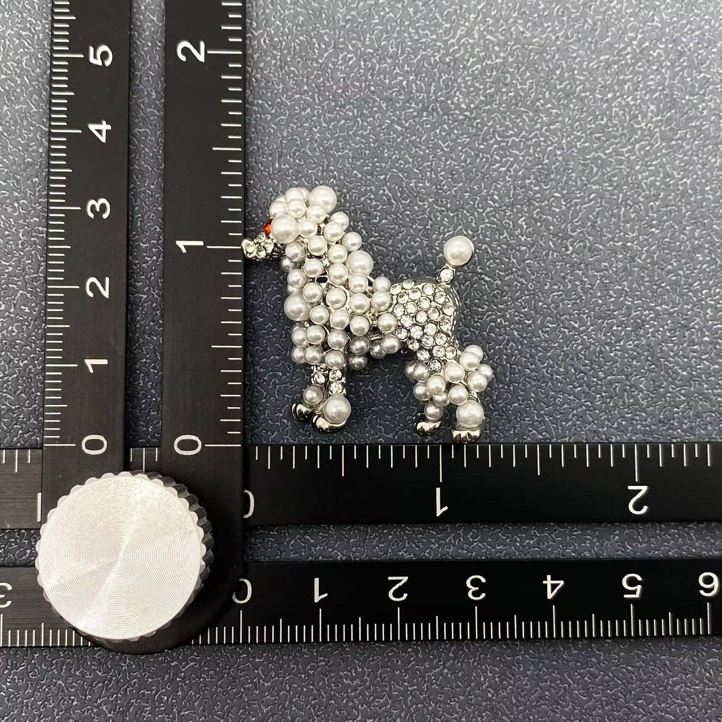 Silver poodle-shaped brooch with pearl and rhinestone embellishment on a textured black surface with a ruler for scale.