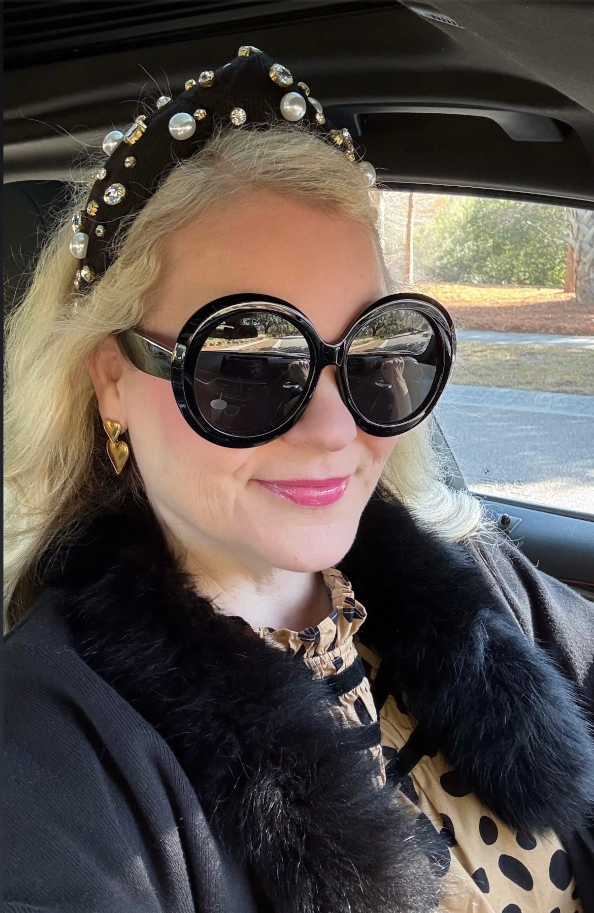 a woman with blonde hair and black headband and black faux fur coat wearing black round sunglasses in a car