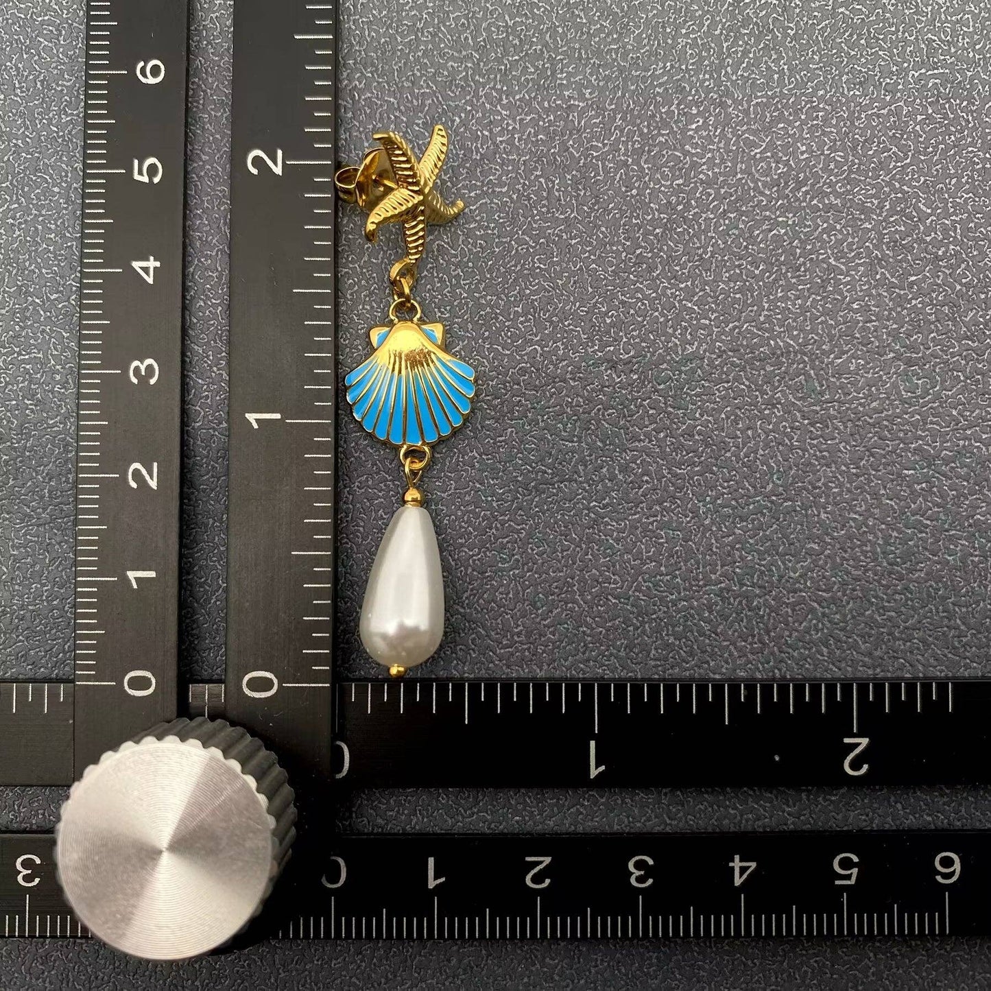 one coastal inspired earring with gold starfish, gold and blue enamel clam shell and pearl drop against a black ruler and grey background