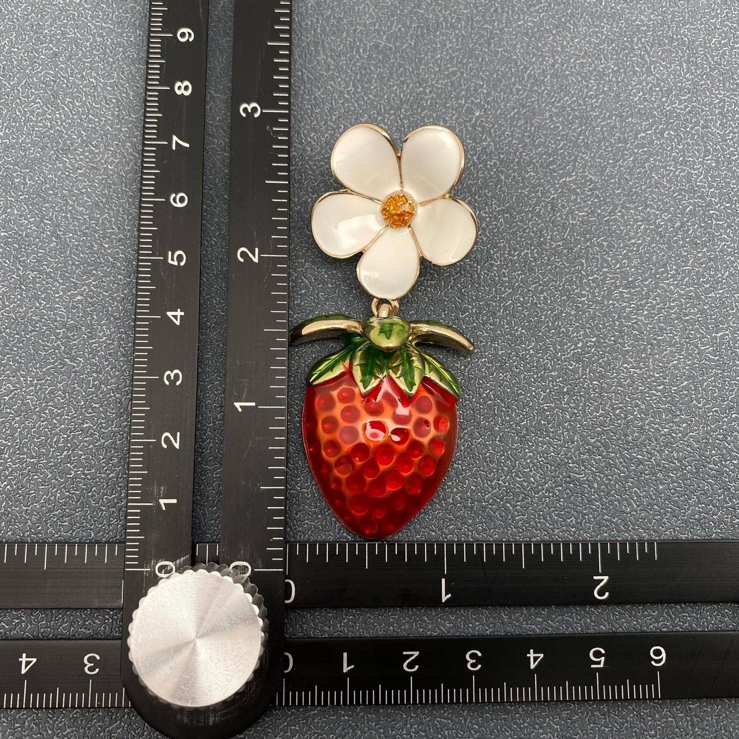a single strawberry and daisy themed enamel earring next to a black ruler on a grey background