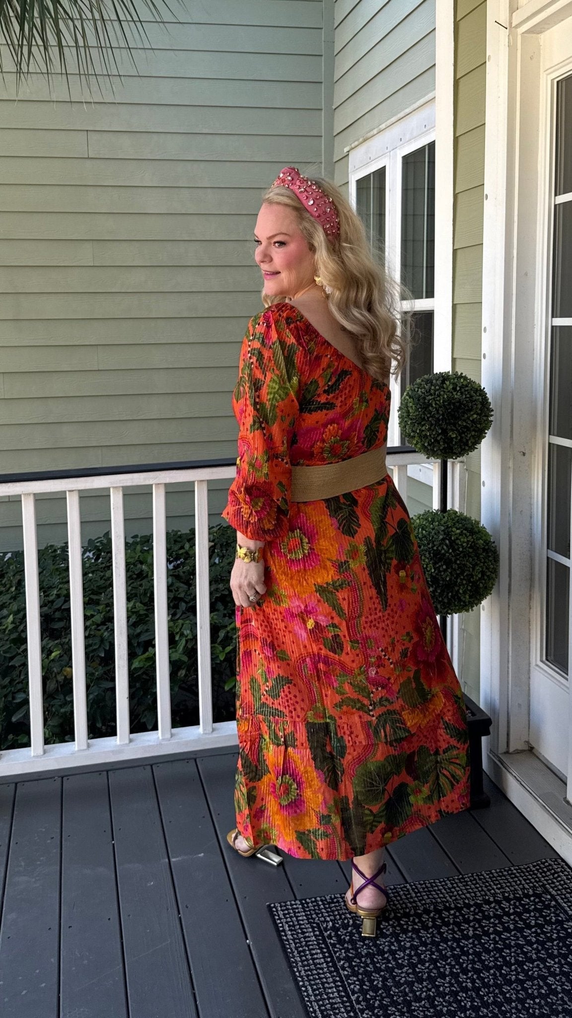 Woman in an orange one-shoulder dress standing on a porch with greenery and a house in the background