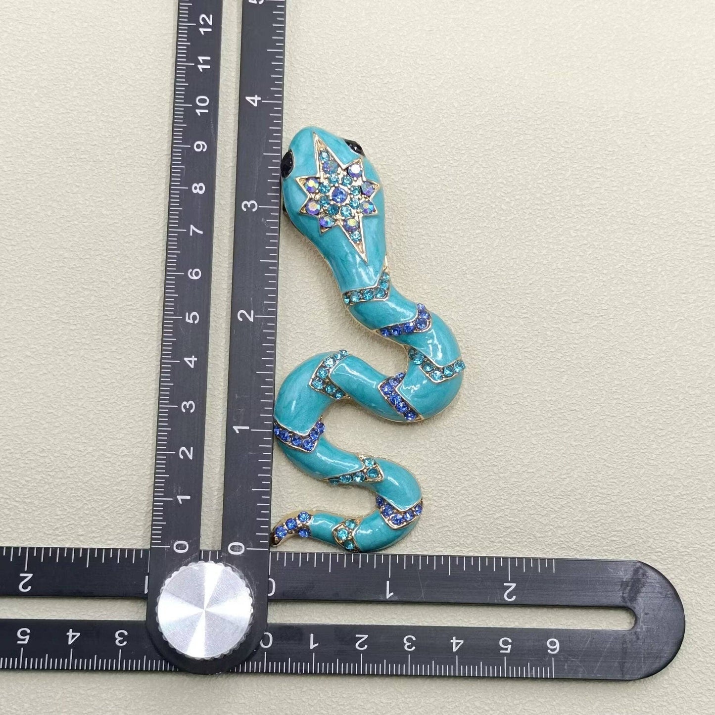 Turquoise snake-shaped decorative brooch next to a ruler on a light gray background