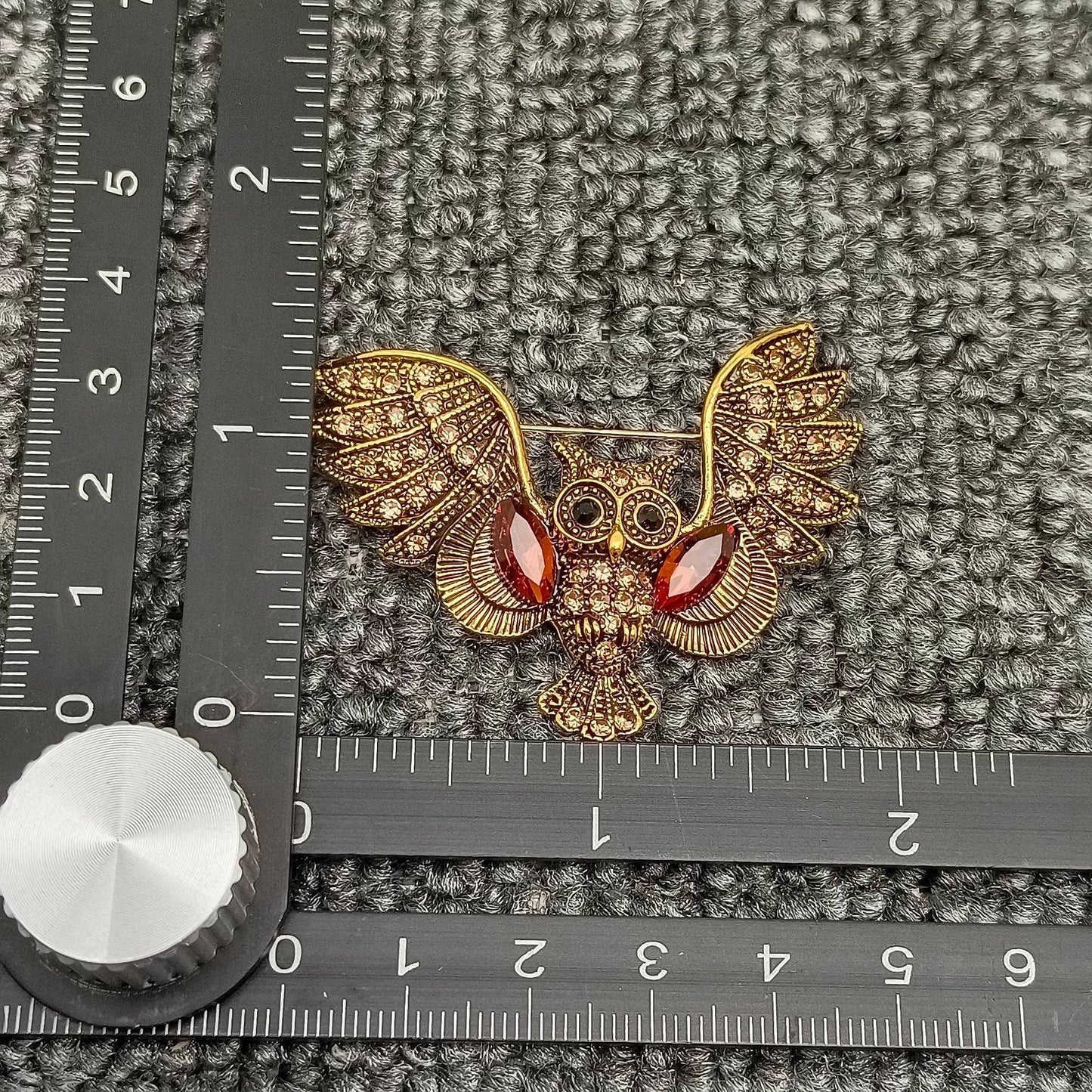 Gold owl brooch with cognac colored gemstones on a grey textured surface with a ruler for scale.