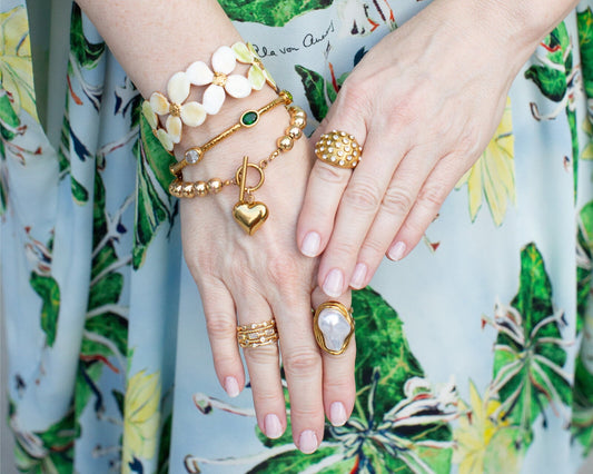 A woman's hands wearing various gold rings and bracelets against a blue and green floral dress