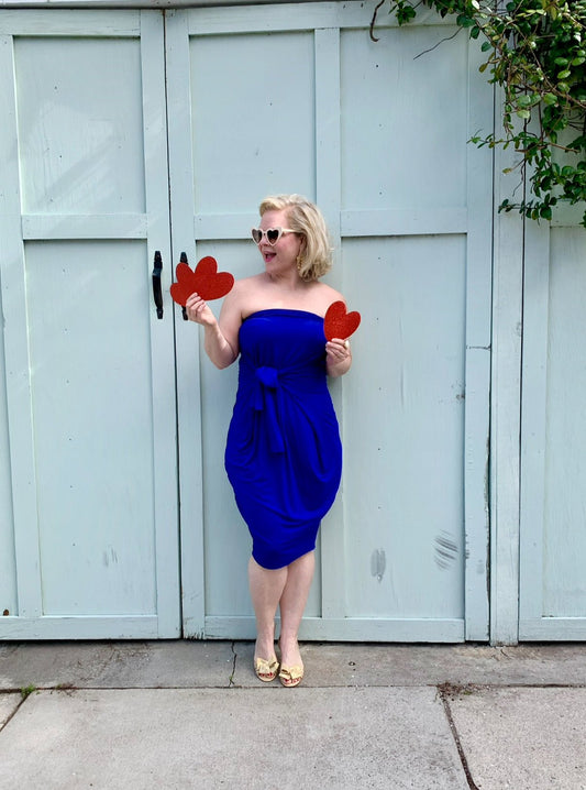 woman in blue dress holding 3 red paper hearts outside in front of a light blue colored garage