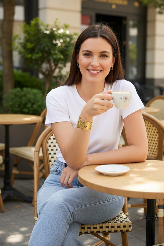 A woman in a white tee and jeans wearing a gold butterfly cuff bracelet while drinking coffee at an outdoor cafe