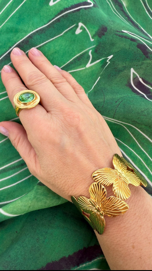 hand wearing a gold and emerald stone cocktail ring and gold butterfly cuff against a green fabric background
