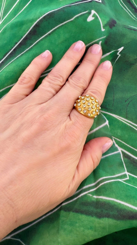 Why Gold jewelry looks good on everyone featuring a gold and crystal cocktail ring on a woman's hand with green and white fabric in the background