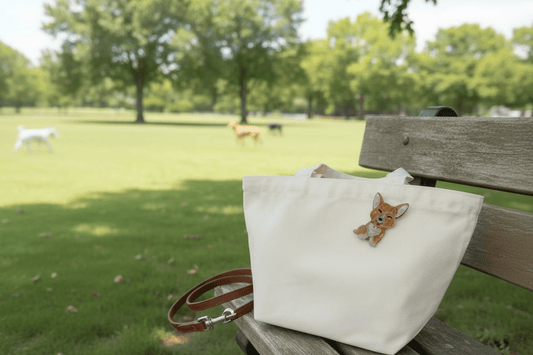 Kitschy Jewelry is back featuring a canvas bag with corgi dog acrylic brooch sitting on a bench at the dog park with dogs in the background