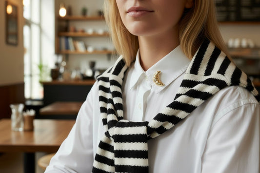 A woman wearing a gold croissant shaped brooch on the collar of a white shirt and a black & white striped sweater around her shoulders in a cafe setting