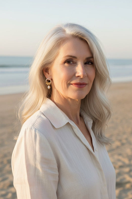 A woman with silver hair wearing gold bow and pearl wreath earrings on a beach with the ocean in the background