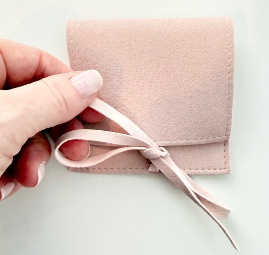 a person's hand tying the ribbon of a pink microsuede jewelry pouch against a light background