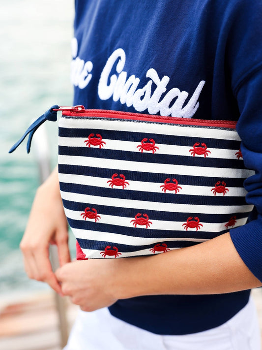 a person carrying a striped crab embroidered pouch and navy shirt outside with the ocean in the background