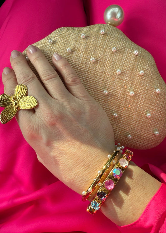 A woman's hand wearing a gold butterfly ring, bracelet, and raffia and pearl encrusted purse against a pink dress