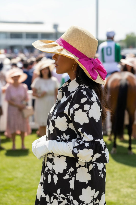 A woman in a raffia and pink bow hat, black and white floral dress, and white gloves at a horse race outside