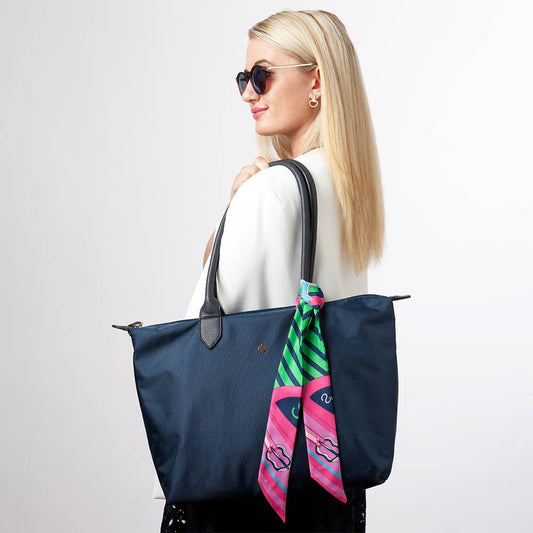 A woman in a white top wearing a navy tote bag with a pink, green, and blue scarf tied to the handle against a white background