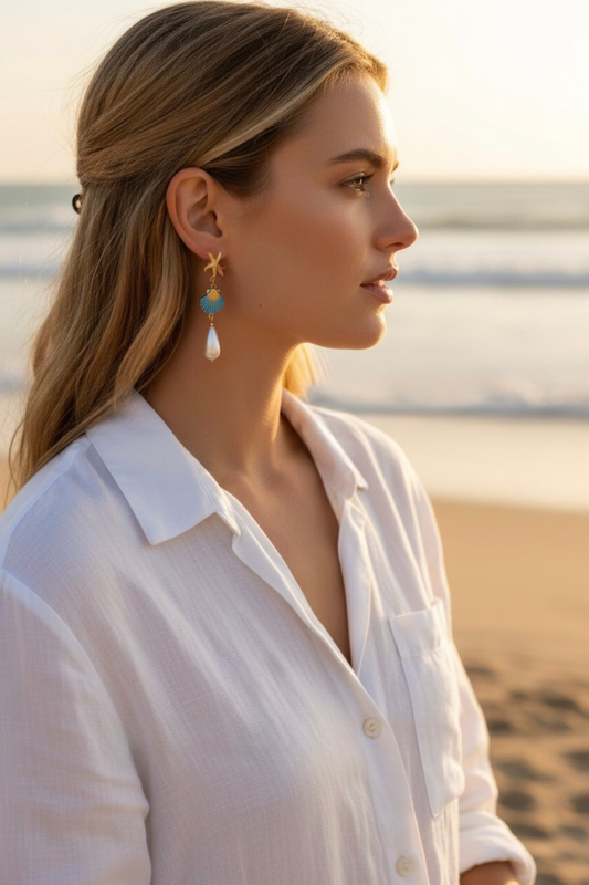 woman in a white blouse wearing a gold starfish and pearl drop earring in a beach setting