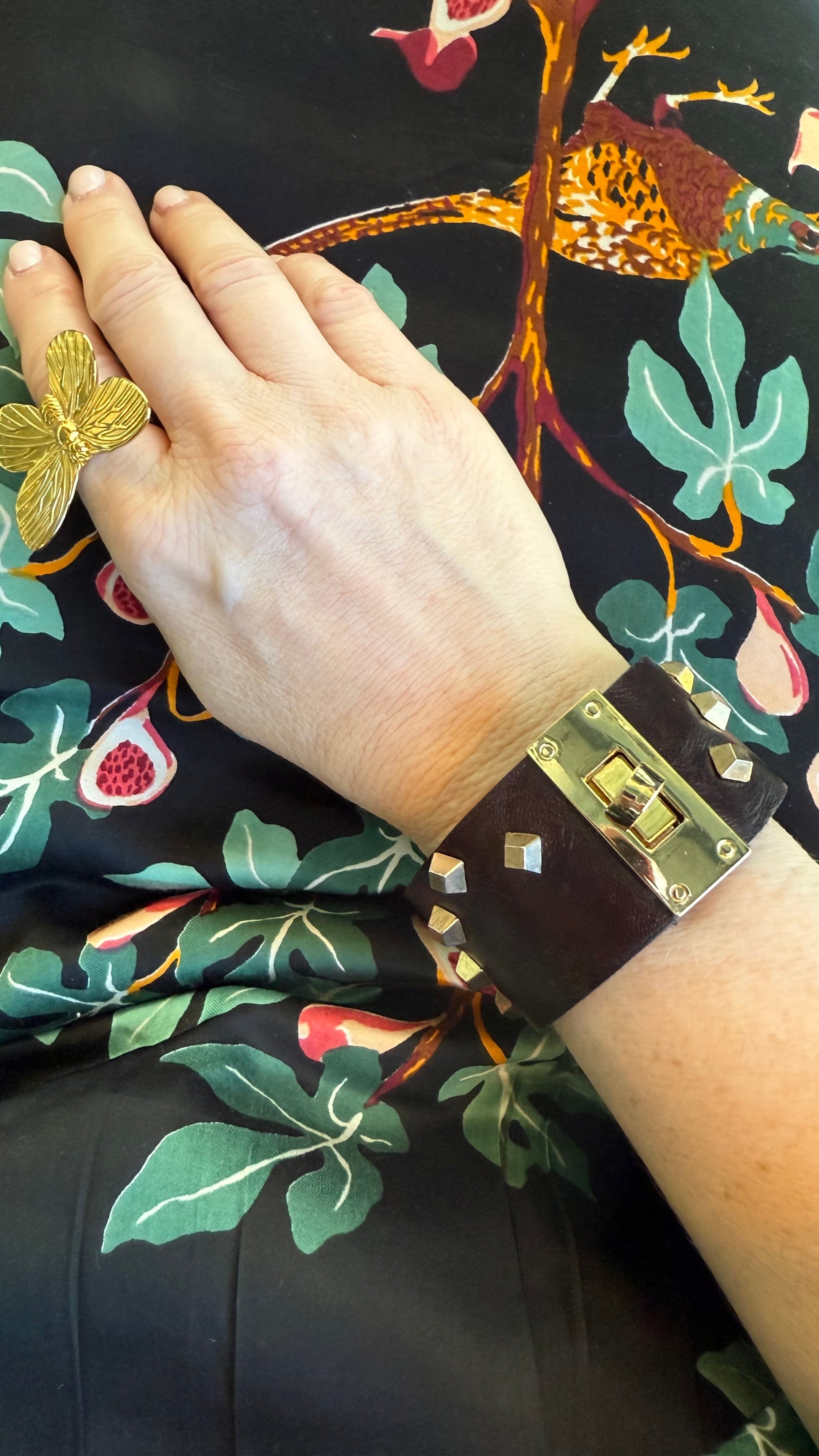 Hand wearing a brown leather and gold studded cuff bracelet on a dark fabric with floral and bird patterns