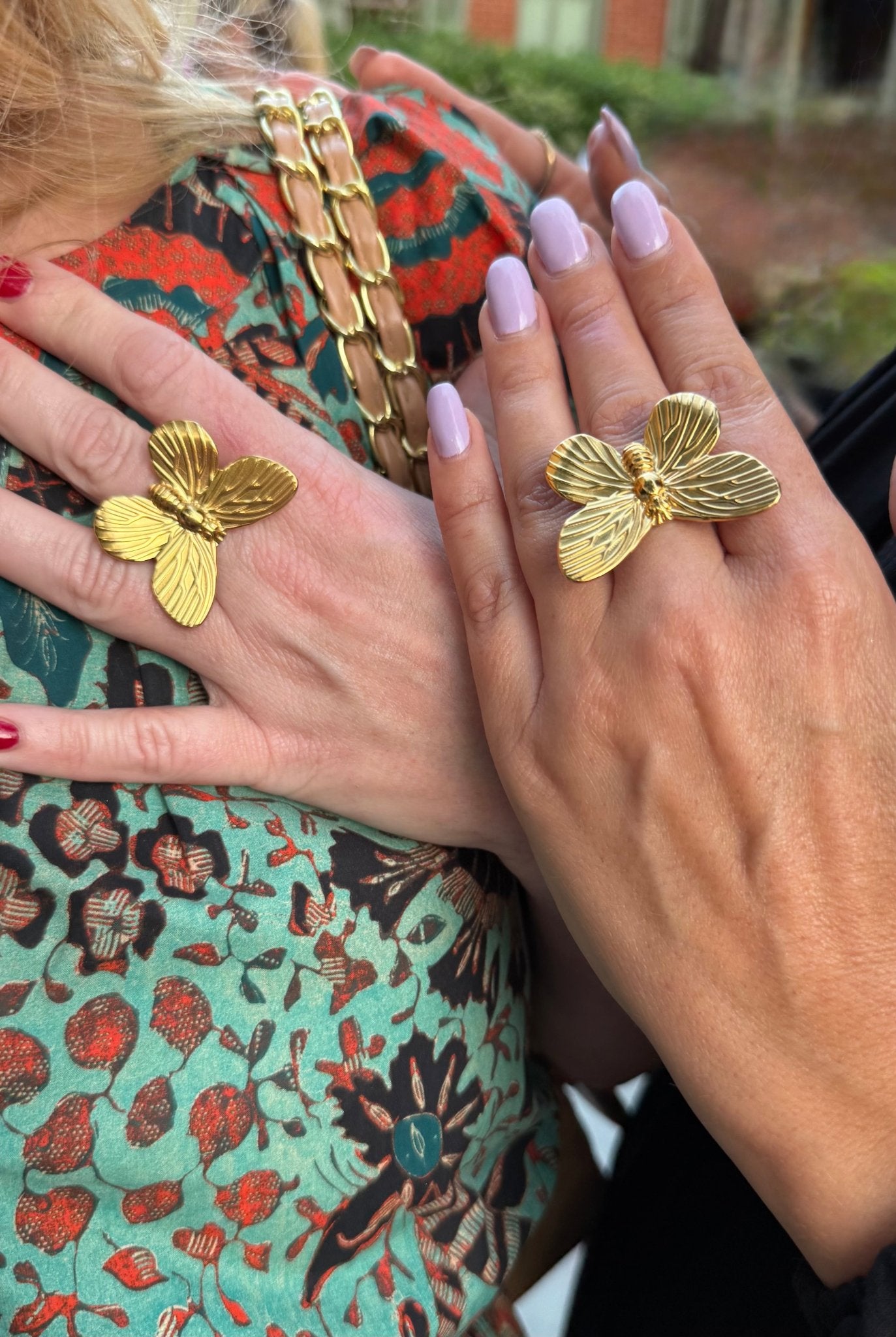 Close-up of hands wearing gold butterfly rings with a colorful patterned garment in the background.