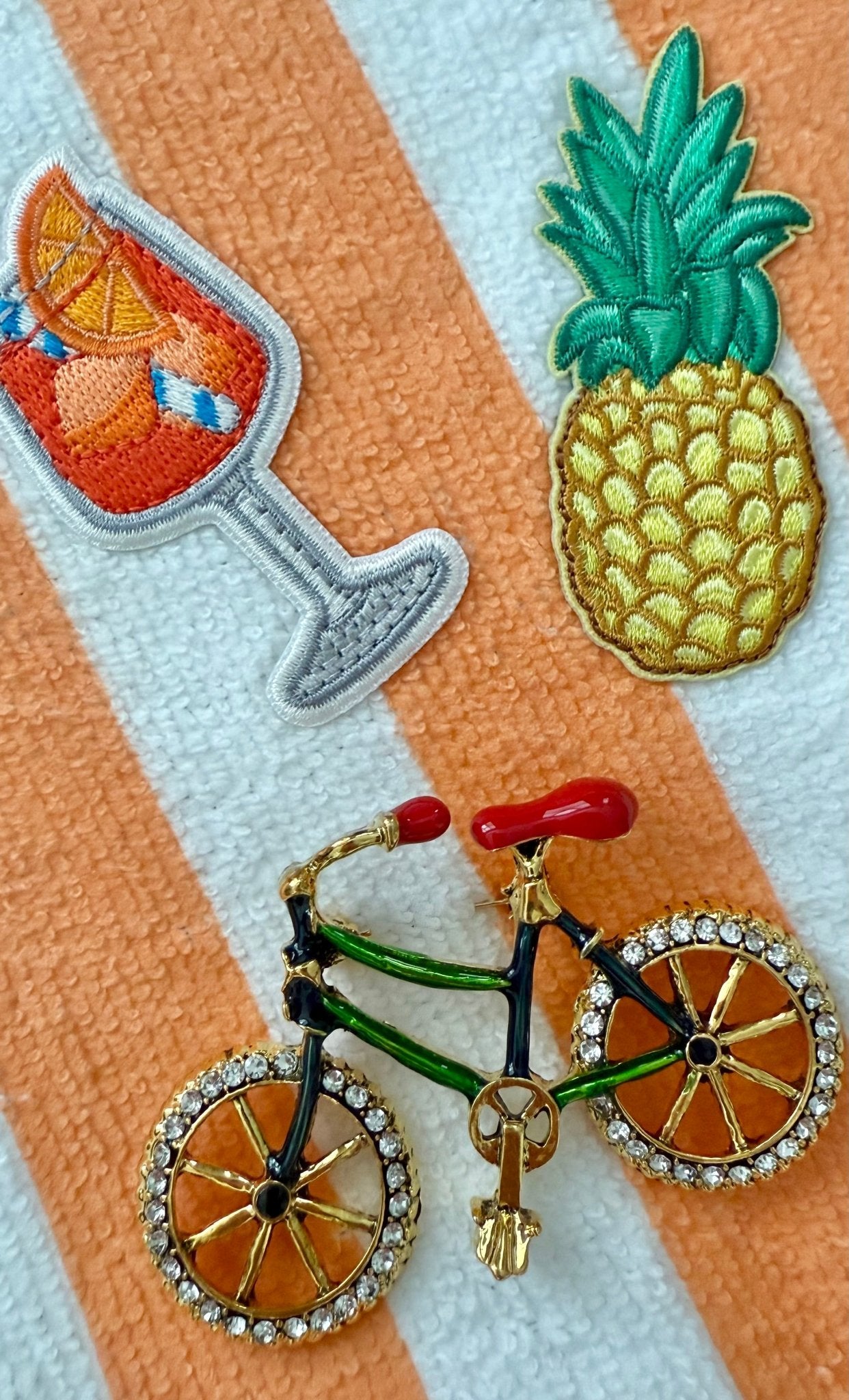 Embroidered patches of a pineapple, and aperol cocktail, and a bicycle brooch on a striped terrycloth background