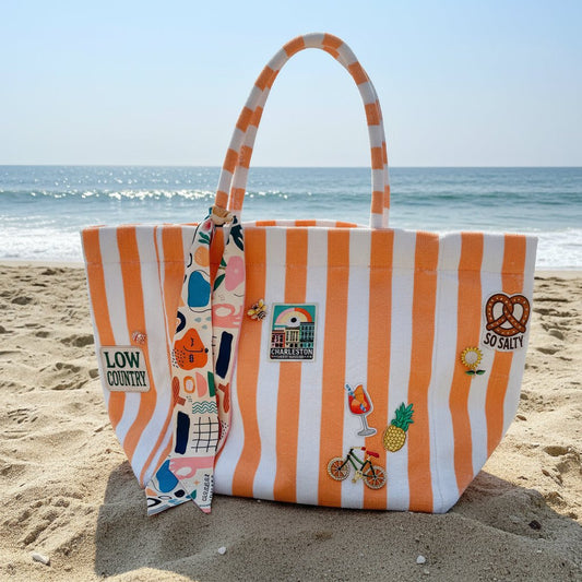 Coral striped terrycloth tote bag with various patches, brooches, and a scarf on the sand at the beach