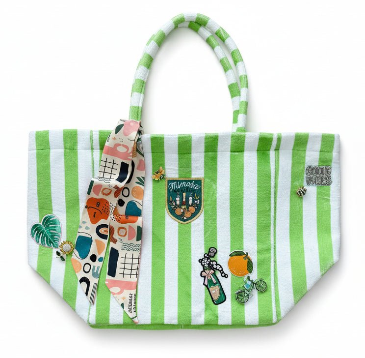 Green and white striped terrycloth tote bag with colorful patches, brooches, and a scarf on a white background