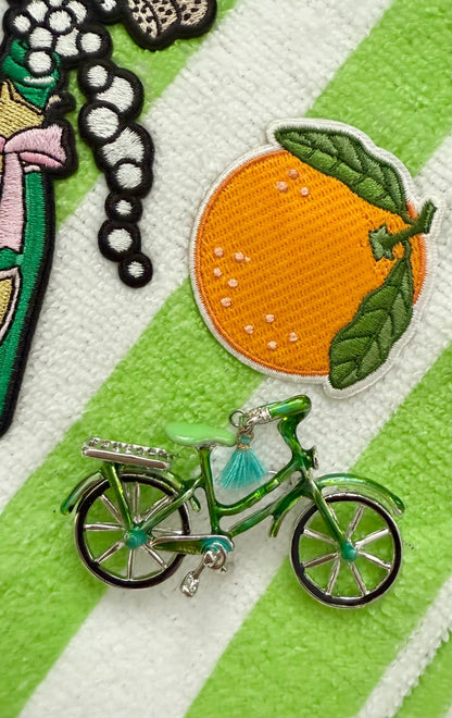 Green bicycle brooch and orange fruit patch on a striped green and white background 