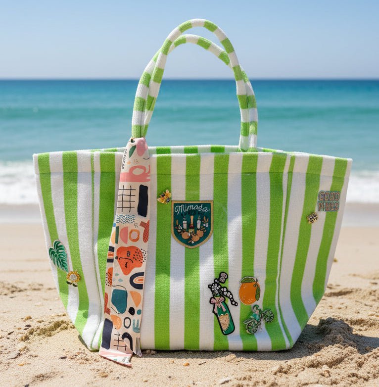 Green and white striped terrycloth tote bag with decorative patches, brooches, and a scarf in a beach setting