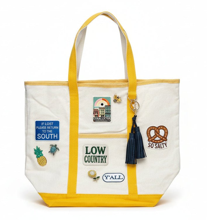 White tote bag with yellow straps and various patches, brooches, and a tassel on a white background
