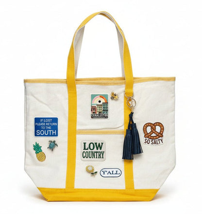 White tote bag with yellow straps and various patches, brooches, and a tassel on a white background