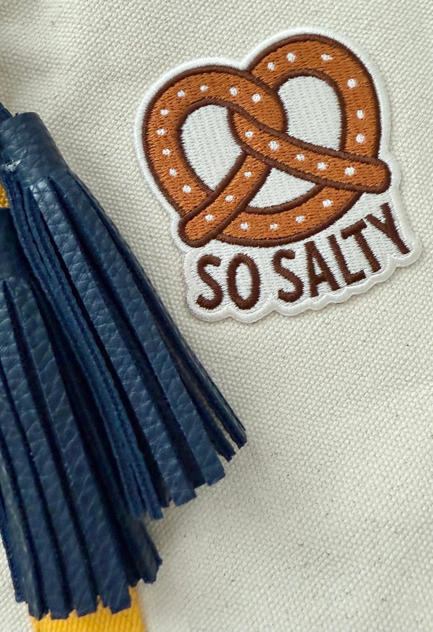 Patch with pretzel design and 'So Salty' text and a blue leather tassel on a canvas fabric background