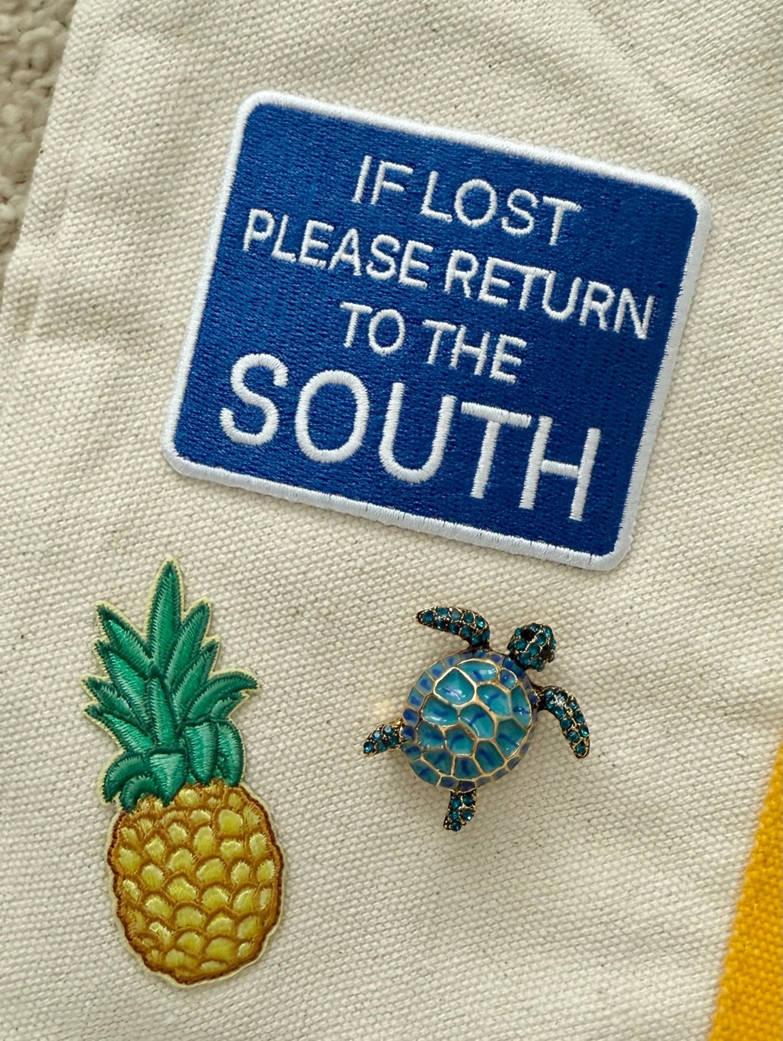 Embroidered patch with pineapple, turtle brooch, and text 'If lost please return to the South' on a canvas fabric background.