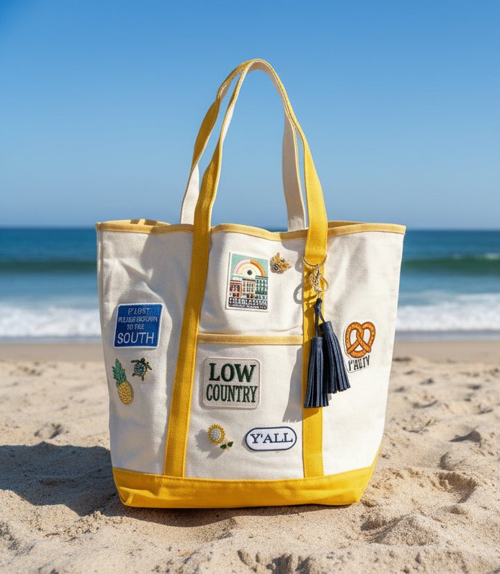 White tote bag with yellow accents and patches and brooches on a sandy beach with ocean in the background