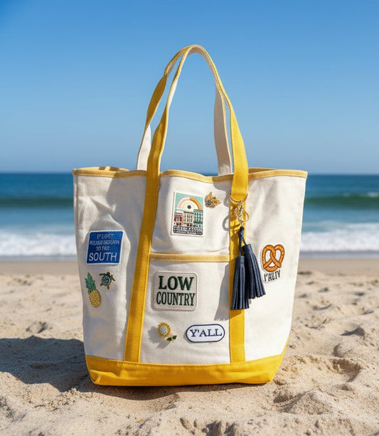 White tote bag with yellow accents and patches and brooches on a sandy beach with ocean in the background