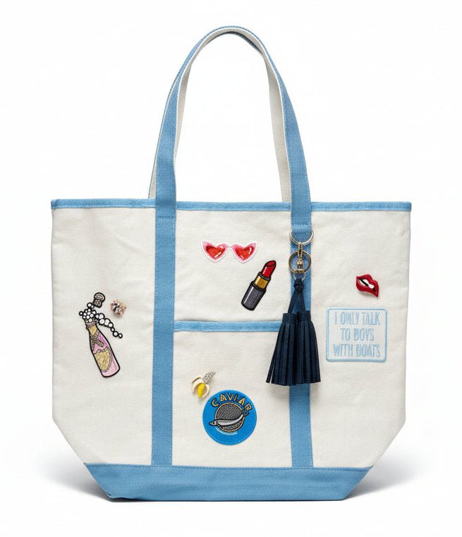 White canvas tote bag with blue straps and various decorative patches and brooches on a white background