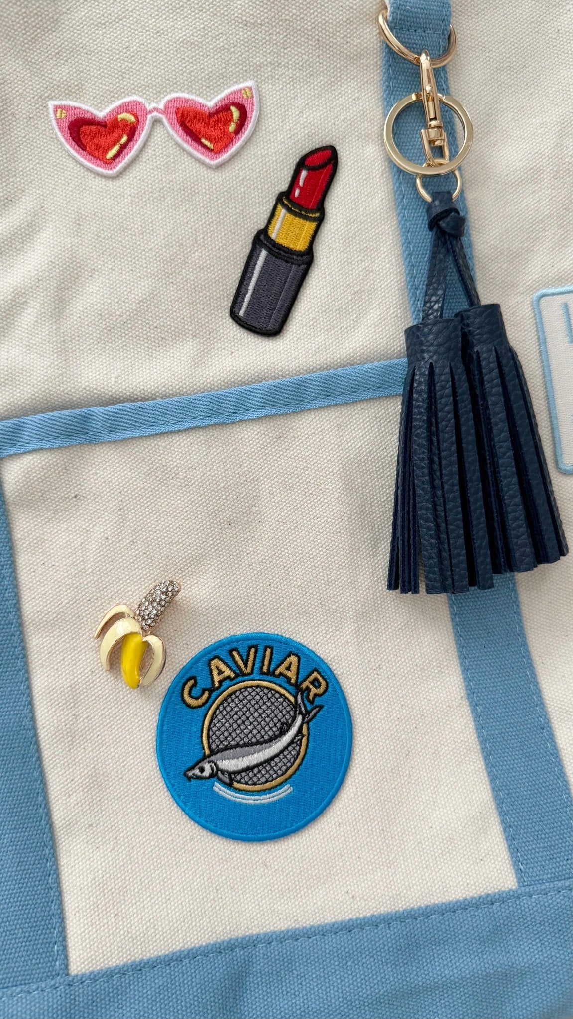 Up close view of blue keychain with tassel, and various patches and banana brooch on an ivory canvas bag with light blue trim