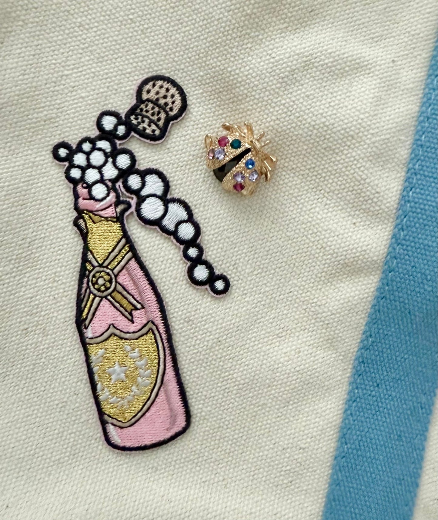 Embroidered patch of a champagne bottle with a lady bug brooch on a canvas fabric background