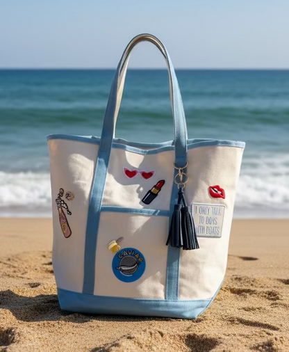 Beige canvas tote bag with blue straps and various patches and brooches on the sand at the beach.