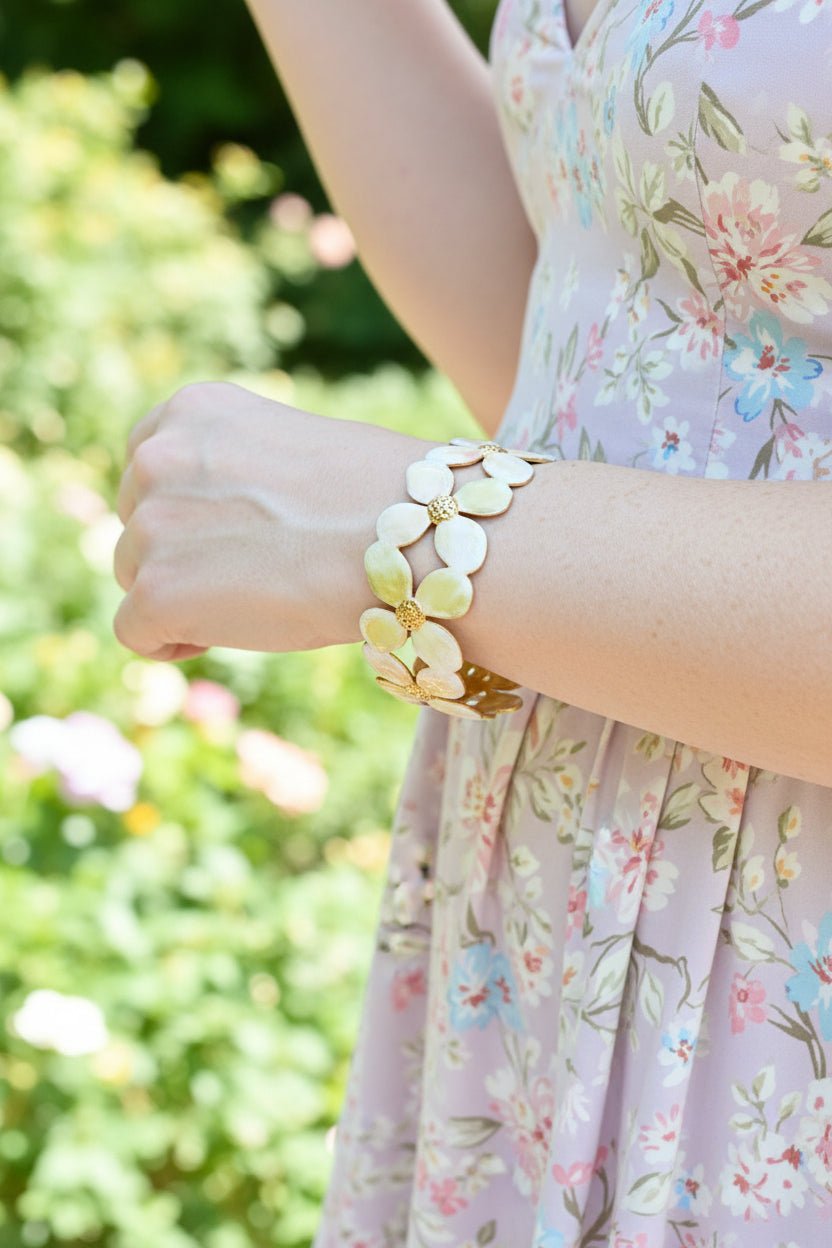 a woman's wrist with a gold and enamel flower cuff bracelet and a floral dress in an outdoor setting.