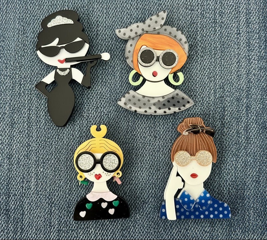 Four decorative acrylic fashionista brooch pins of stylized women with unique hairstyles and accessories on a denim background.