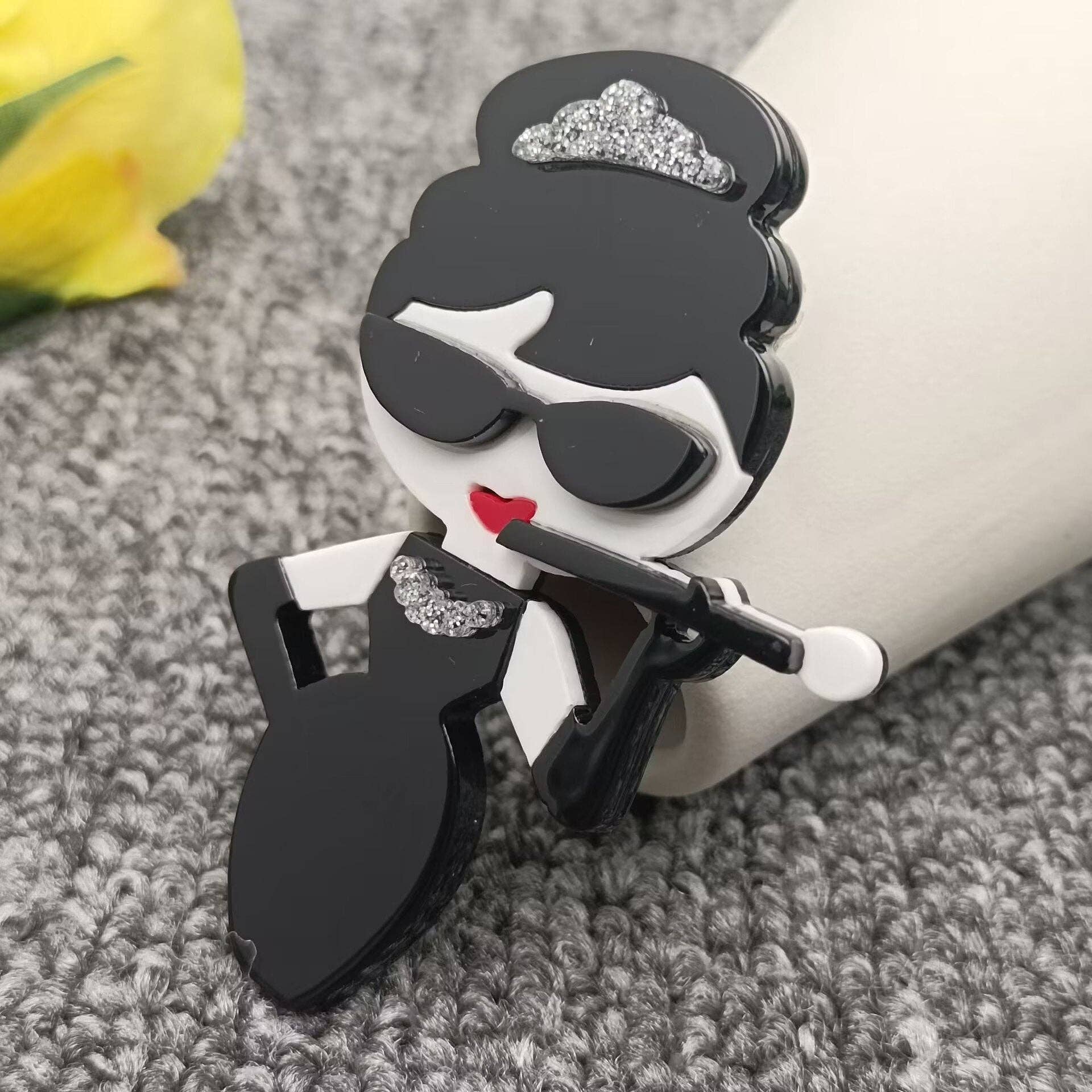 Audrey fashionista brooch on a grey carpet background leaning against a white vase.