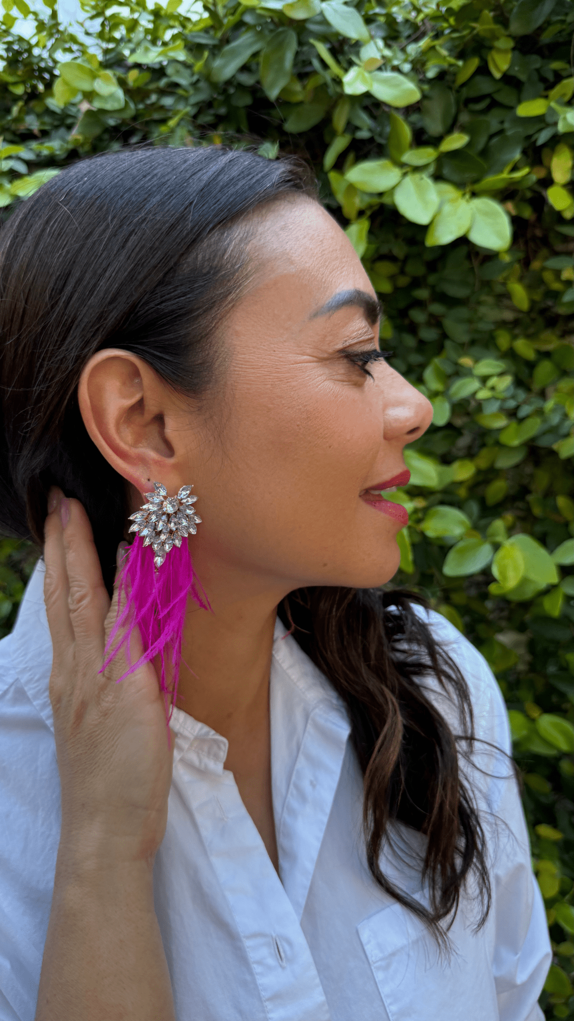 Woman wearing a white shirt and pink fuchsia feather earrings with a blurred green background
